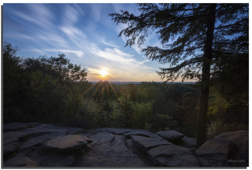 The sun is setting over the horizon at Ledges Trail Overlook. The sun reflects a golden hue on the trees.