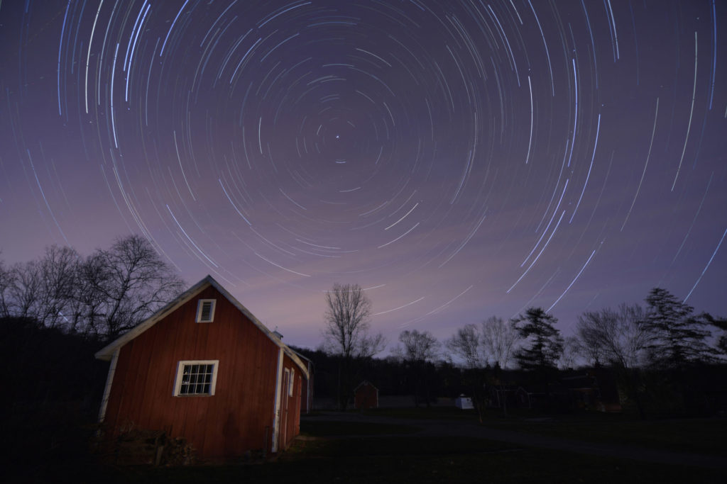 A long-exposure image taken at night shows a circle of stars rotating above a red barn.