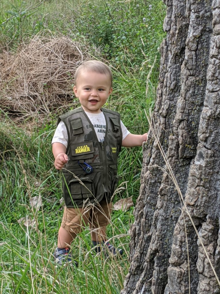 Little boy wearing Jr. Ranger vest plays peek-a-boo by tree