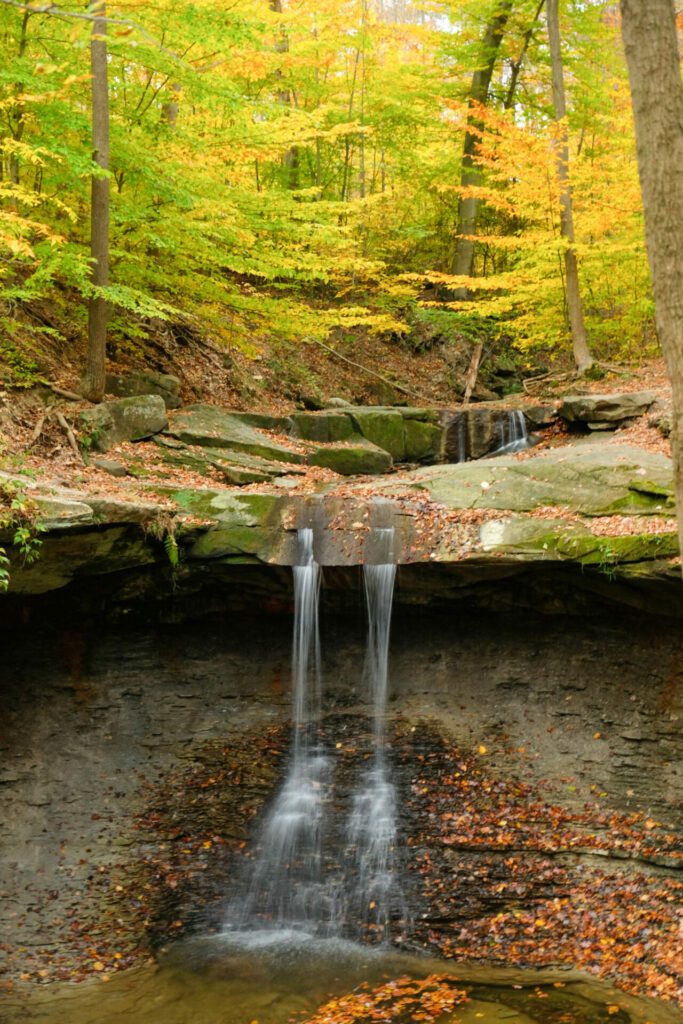 The image is a photograph of Blue Hen Falls in Cuyahoga Valley National Park. The photo shows a waterfall cascading over a natural stone ledge covered with green moss in some spots. The trees in the background have green leaves but some leaves have turned yellow or orange. 