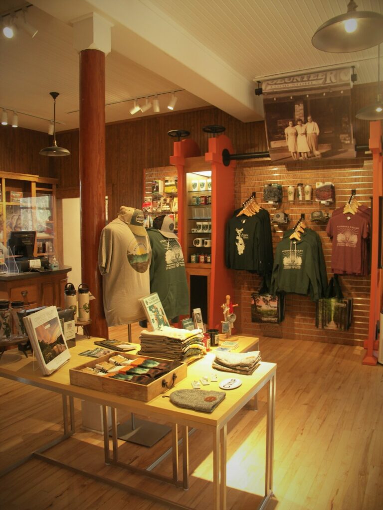 Two display tables in the Boston Mill Visitor Center gift shop display park-themed items for sale such as a calendar, socks, hats, t-shirts, and mugs. Across from the tables is a display wall with hanging merchandise such as sweatshirts, t-shirts, and tote bags. 