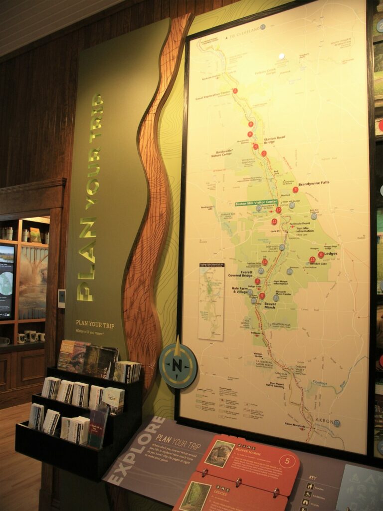 Wall signage reads Plan Your Trip. Next to the signage us a large map of the park with markers for popular attractions and trails. Next to the map is a tiered display holder for free take-along park maps and brochures.