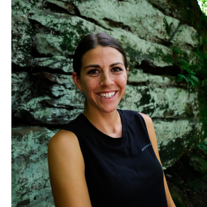 A smiling woman in a black sleeveless t-shirt standing in front of a rock wall covered in light green lichens. 