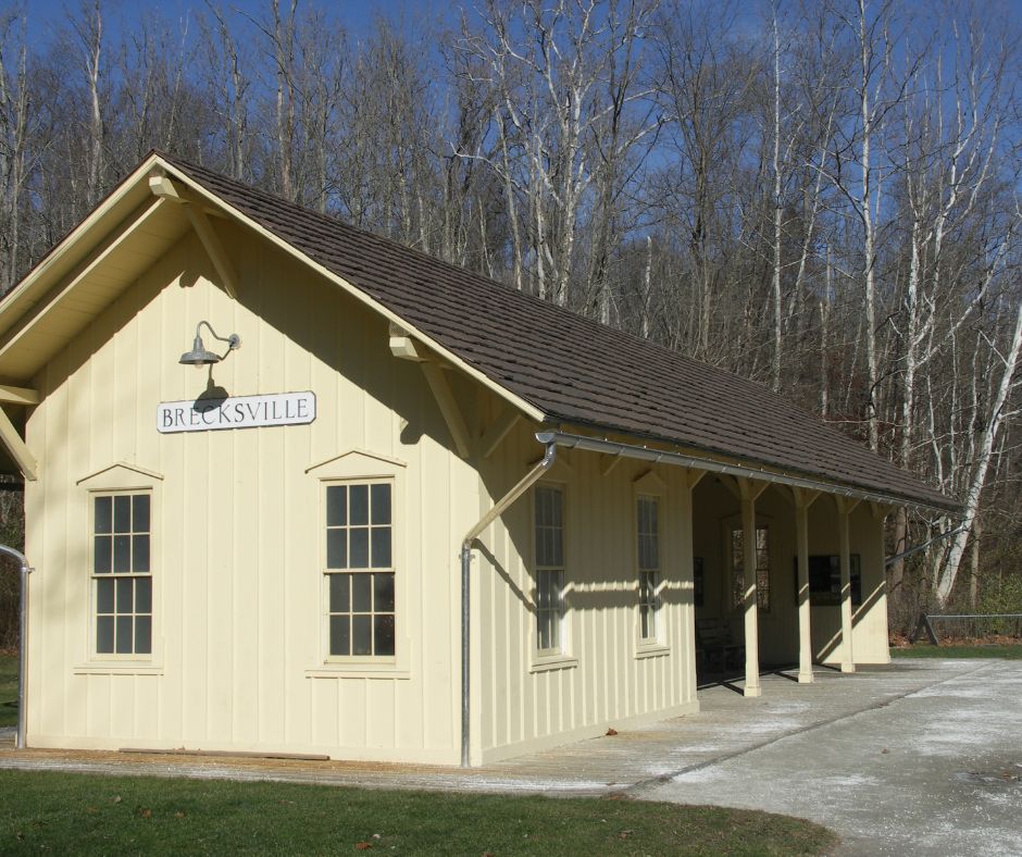 The Brecksville train station is shown in the foreground. It is a pale-yellow building with a peaked, shingled roof. From the front, it resembles a small one-story house with windows, but part of the structure is open. Under the roof of the open section, train passengers can wait. In the background bare trees stand tall against the bright, blue sky.