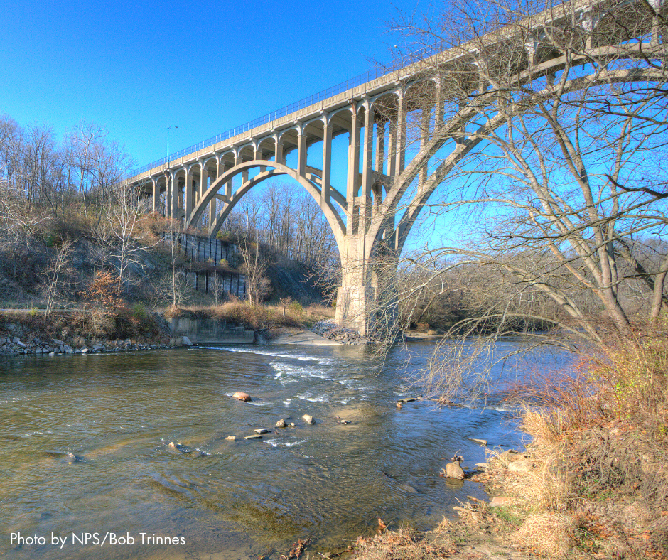 The two arches of the tall bridge that carries Route 82 through the valley is shown at an angle. Underneath the arches is the Cuyahoga River. Rocks can be seen in the river and along the banks. Bare trees and thick brush are part of the landscape. The sky is bright blue.
