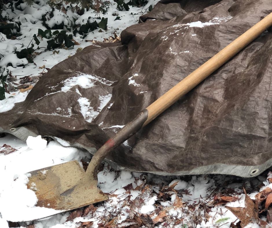 A compost pile is covered by a brown, plastic tarp. A long-handled shovel rests across the tarp. The ground by the compost pile is covered in brown, dried leaves and snow. 