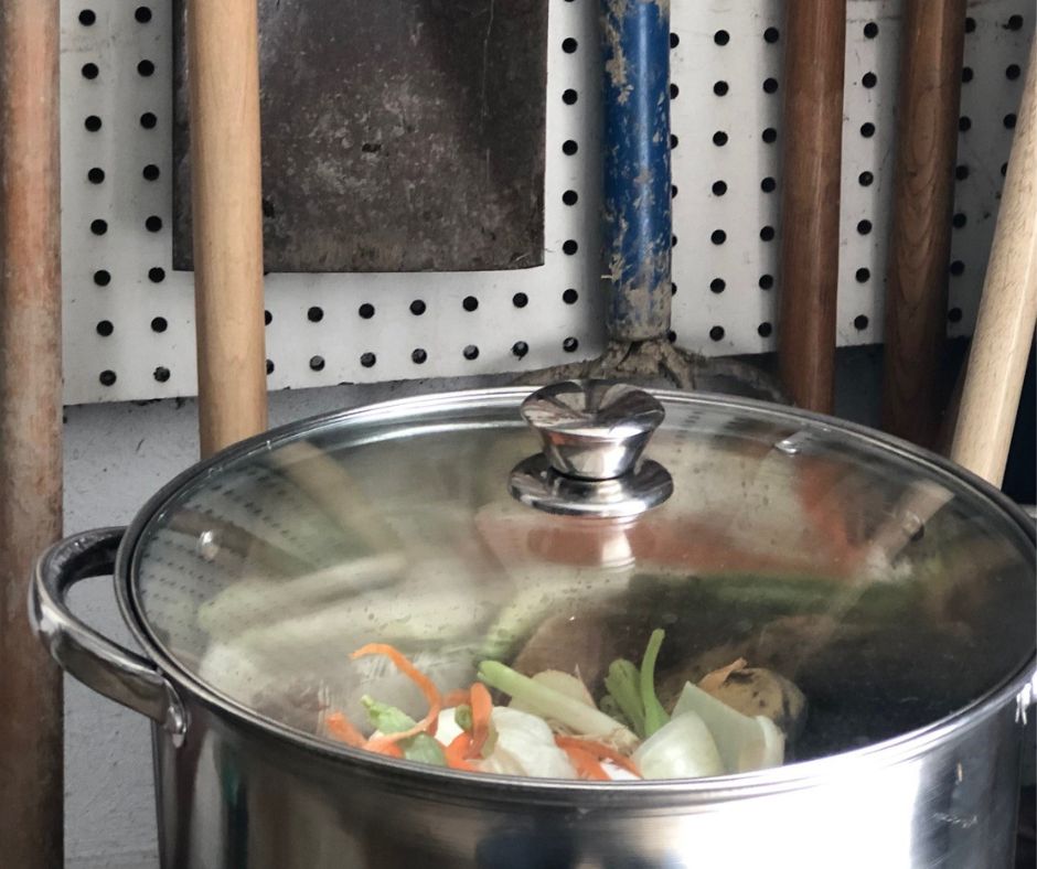 The shot is taken in the interior of house garage. A 16-gallon stainless steel stock pot sits in the foreground. The lid of the stock pot is clear. Food scraps such as celery stalks, carrot shavings, and onion peel are within the pot. Behind the pot is a pegboard wall. Several garden tools hang from the pegboard.