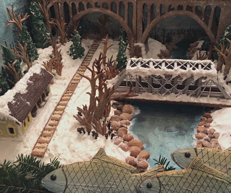 A gingerbread landscape of the Station Road area depicts the Station Road Bridge and the Brecksville Train Station. The scene includes railroad tracks by the train station and a blue isomalt version of the Cuyahoga River. In the back of the scene is a gingerbread version of the arched bridge that carries Route 82 through the valley. The landscape is iced to look snowy and has trees and plants made from gingerbread and green chocolate. In the foreground are three fish-shaped cookies that represent bigmouth buffalo fish. The fish are silvery-blue, have teched scales, and dark, circular eyes. 