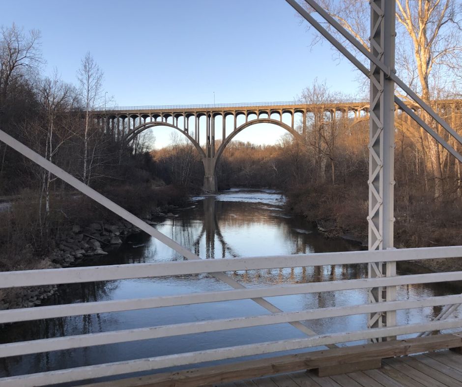 The image depicts a view from the historic Station Road Bridge of the Cuyahoga River and another bridge that carries Route 82 through the valley. Some of the wrought iron beams and rails of the Station Road Bridge are in the foreground. The other bridge in the distance is tall and is comprised of several elegant arches. The Cuyahoga River runs beneath both bridges. A train track can be seen near the rocky left bank of the river. Bare trees and dense brush are on either side of the river and in the distant landscape. 