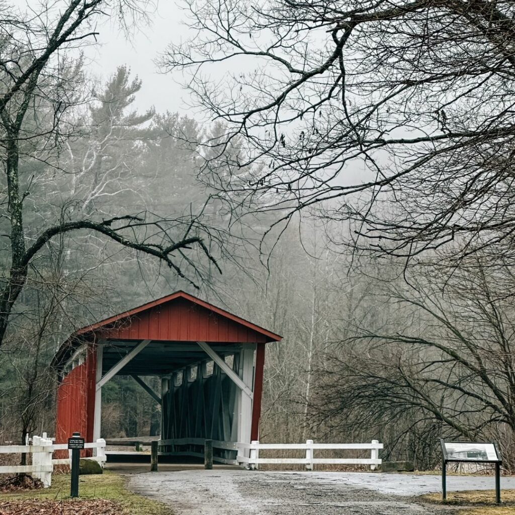 A covered bridge painted in a rusty red color with white inner beams. The day is gray and foggy and bare trees are in the landscape. A gravel path leads to the entrance of the bridge. 