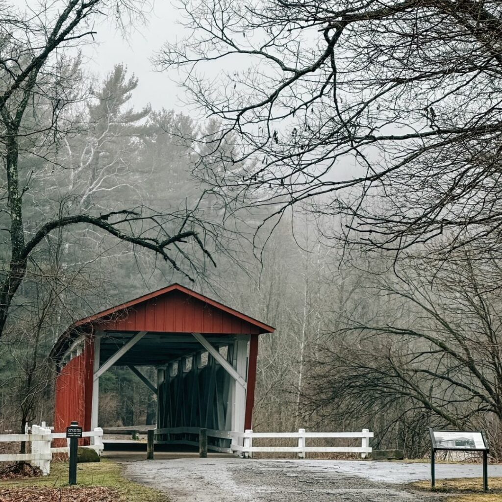 A covered bridge painted in a rusty red color with white inner beams. The day is gray and foggy and bare trees are in the landscape. A gravel path leads to the entrance of the bridge.
