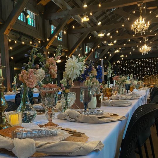 A long table covered in a white tablecloth in a rustic lodge with string lights hanging above it. The table is set with dishes, cloth napkins, candlesticks, votives, wine glasses, and flowers in vases.