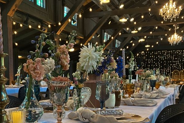 A long table covered in a white tablecloth in a rustic lodge with string lights hanging above it. The table is set with dishes, cloth napkins, candlesticks, votives, wine glasses, and flowers in vases.