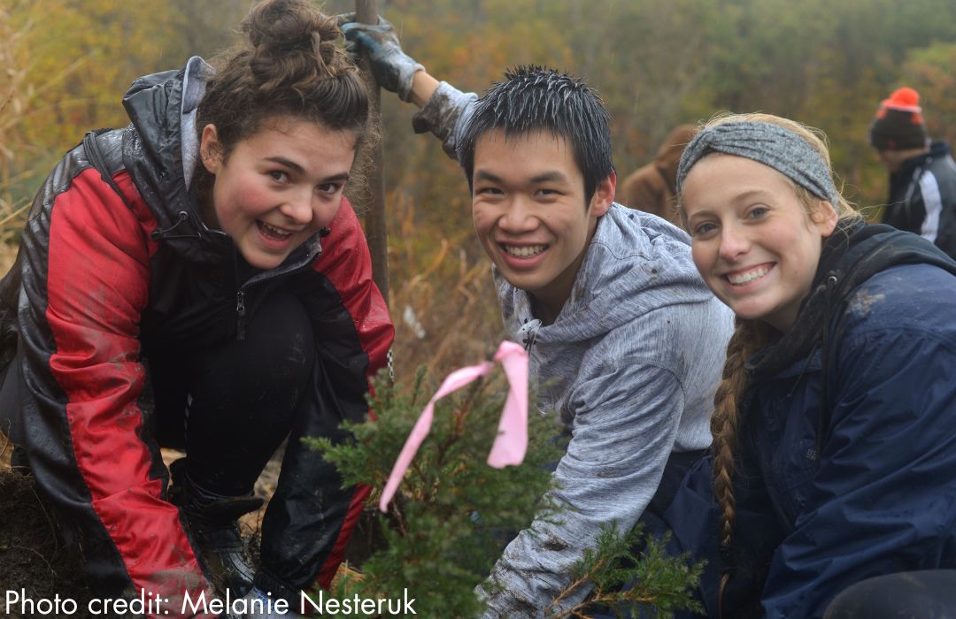 Three volunteers are kneeling on the ground and smiling as they pull plants.