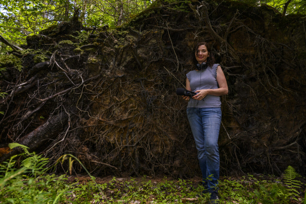 Artist Nikki Lindt stands in a forest. She is holding equipment for recording sound and has headphones around her neck.