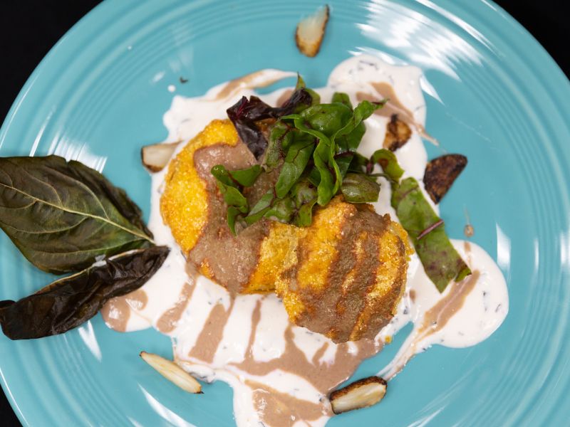 A photo of a turquoise plate with fried green tomatoes stacked on it. The tomatoes are placed in a cream sauce and drizzled with a tan puree. Greens garnish the dish.