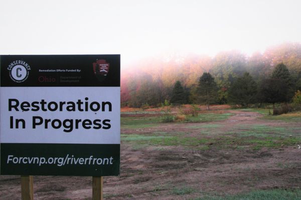 Sign stating restoration in progress in forefront of open field with trees in background.