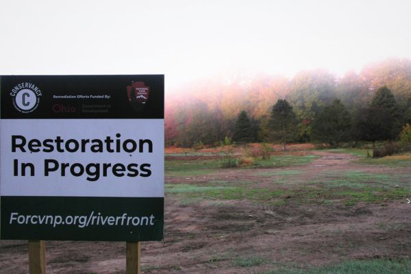 Sign stating restoration in progress in forefront of open field with trees in background.