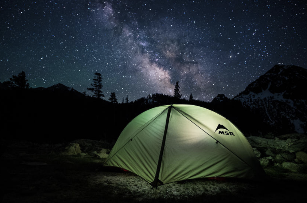 A photo of a camping tent illuminated from the inside. The tent is situated among rocks. Dark trees and a snowy, mountainous landscape are in the background, and a dark, star-filled sky is above. 