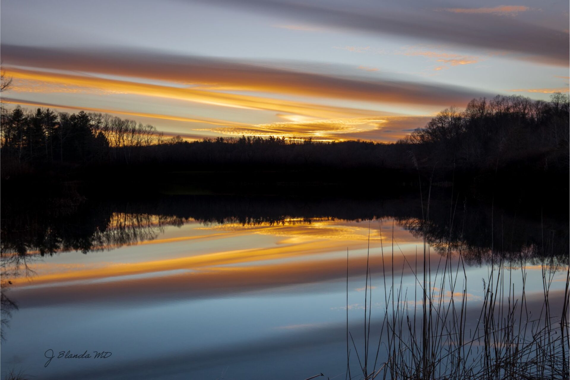 A photo of a pond reflecting the orange light of a sunset, clouds, and blue sky above it. Dark tree silhouettes surround the pond.