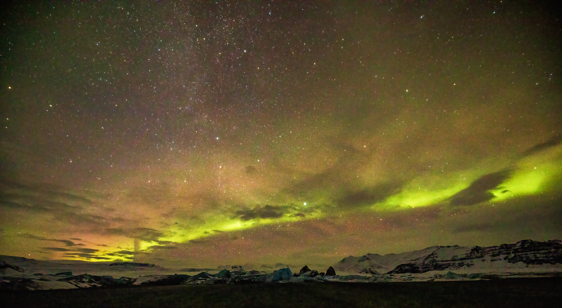 A photo of aurora borealis lights and many stars above a hilly snow-covered landscape. The sky appears to have pink, gray, and lime green hues. 