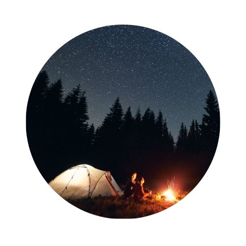 A circular photo of a campsite with a tent illuminated from the inside, a campfire, two people sitting by the fire, dark evergreen tree silhouettes, and a starry night sky. 