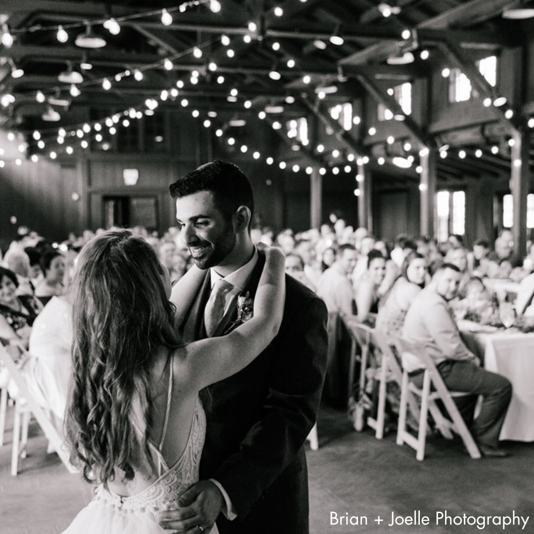 A black and white photo of a bride and groom dancing beneath string lights in a rustic lodge. Weddding guests seated at tables watch.