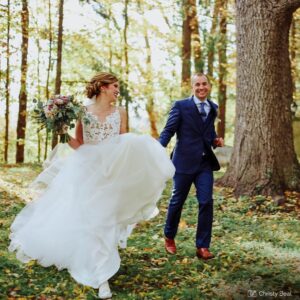 A bride and groom run outdoors, smiling and looking toward each other. Trees are in the background and some fall leaves are on the ground.