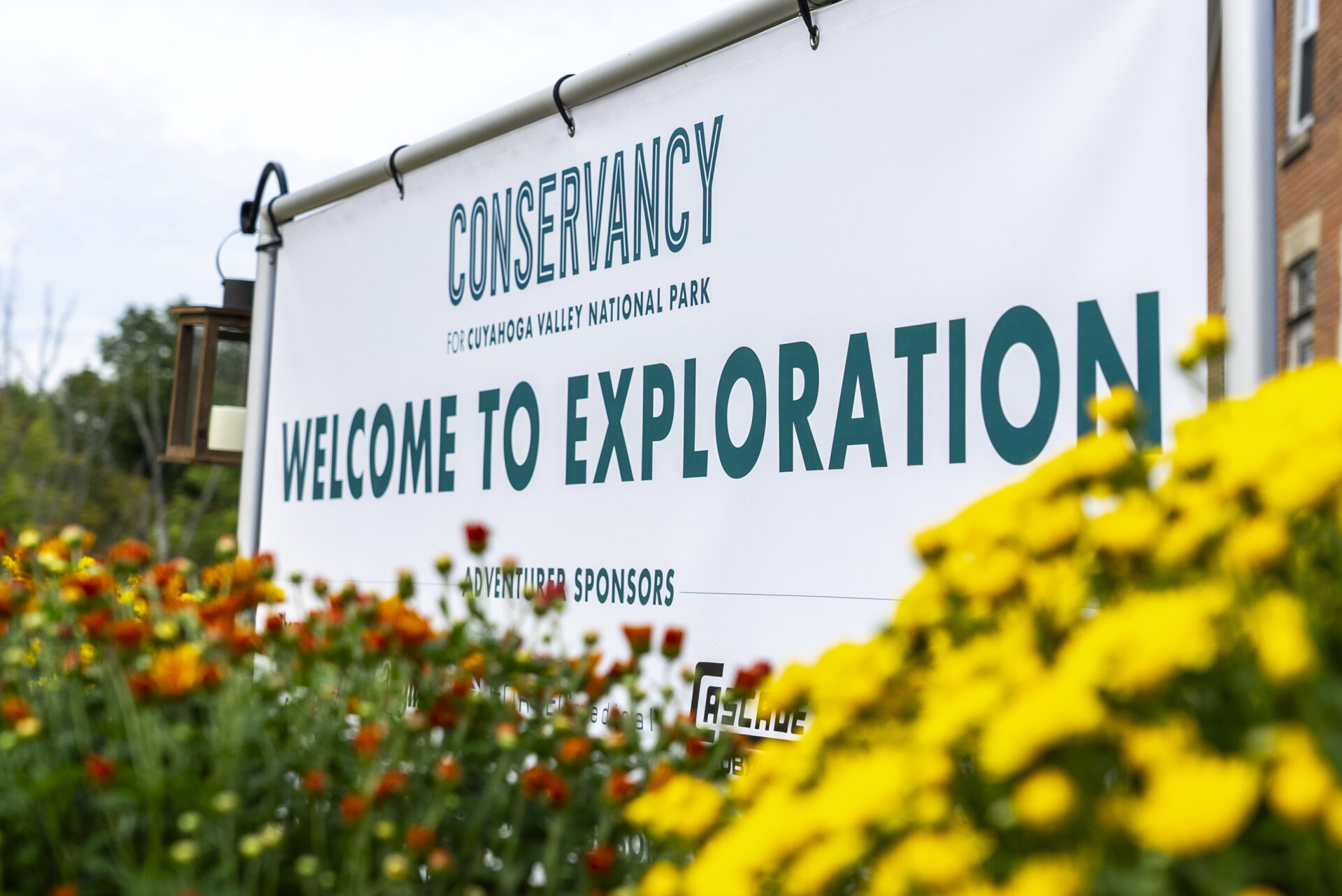 A photo of a sign that includes a Conservancy for Cuyahoga Valley National Park logo and the words: Welcome to Exploration. Fall flowers are in front of the sign and are in hues of orange and yellow.