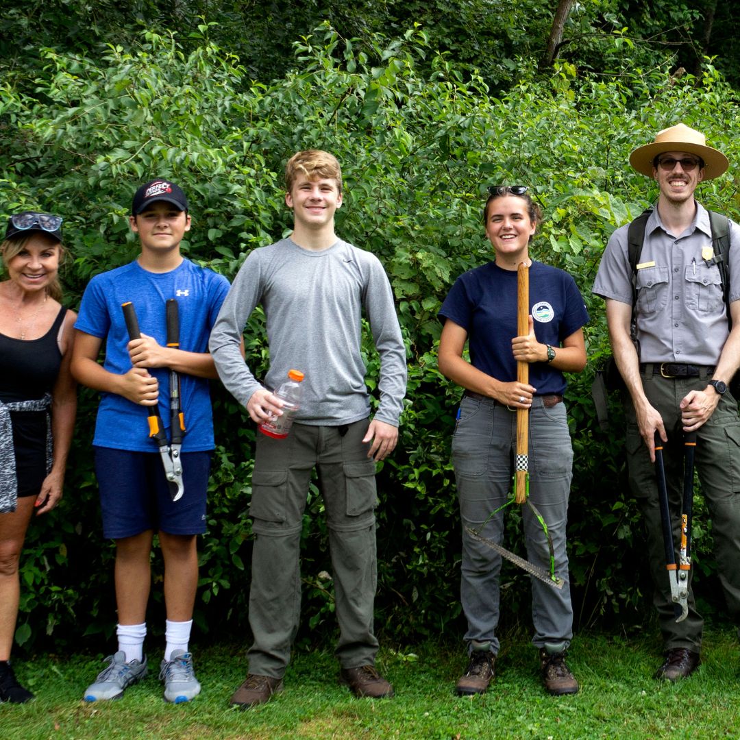 A photo of several people holding hand tools, lined up in front of a hedge in Cuyahoga Valley National Park. One of the people is an NPS ranger in uniform.