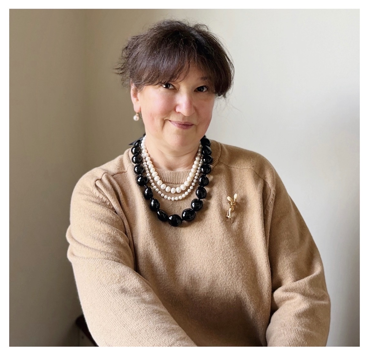 A woman with short dark hair in a tan sweater with chunky necklaces